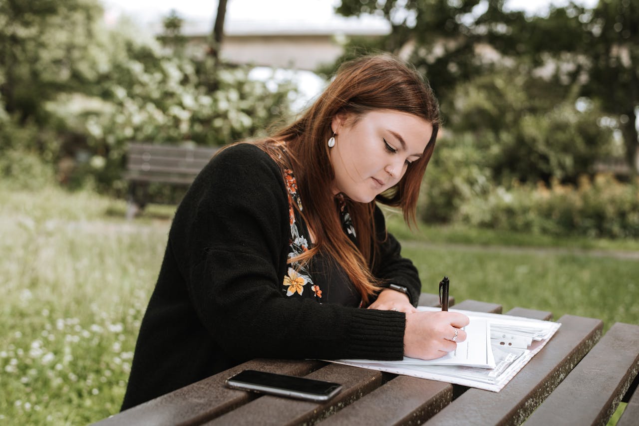 gallery-1 Focused young woman writing in a notebook while studying outdoors in a summer park.
